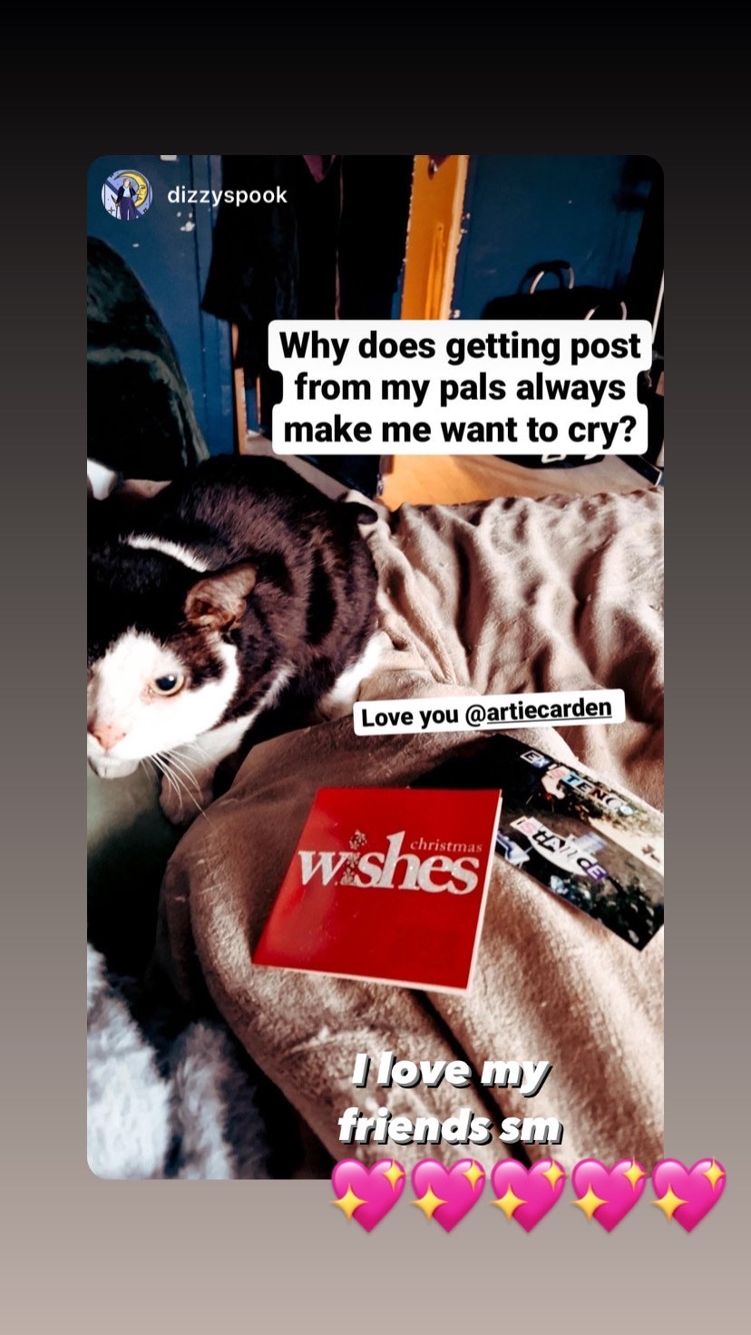 Ig story that shows DizzySpook's lap in bed, her black and white cat on the left and a christmas card and art piece on the bottom right. text reads 'why does getting post from my pals always make me want to cry' from DizzySpook, Artie shared and added 'i love my friends SM' with 5 sparkly pink heart emojis